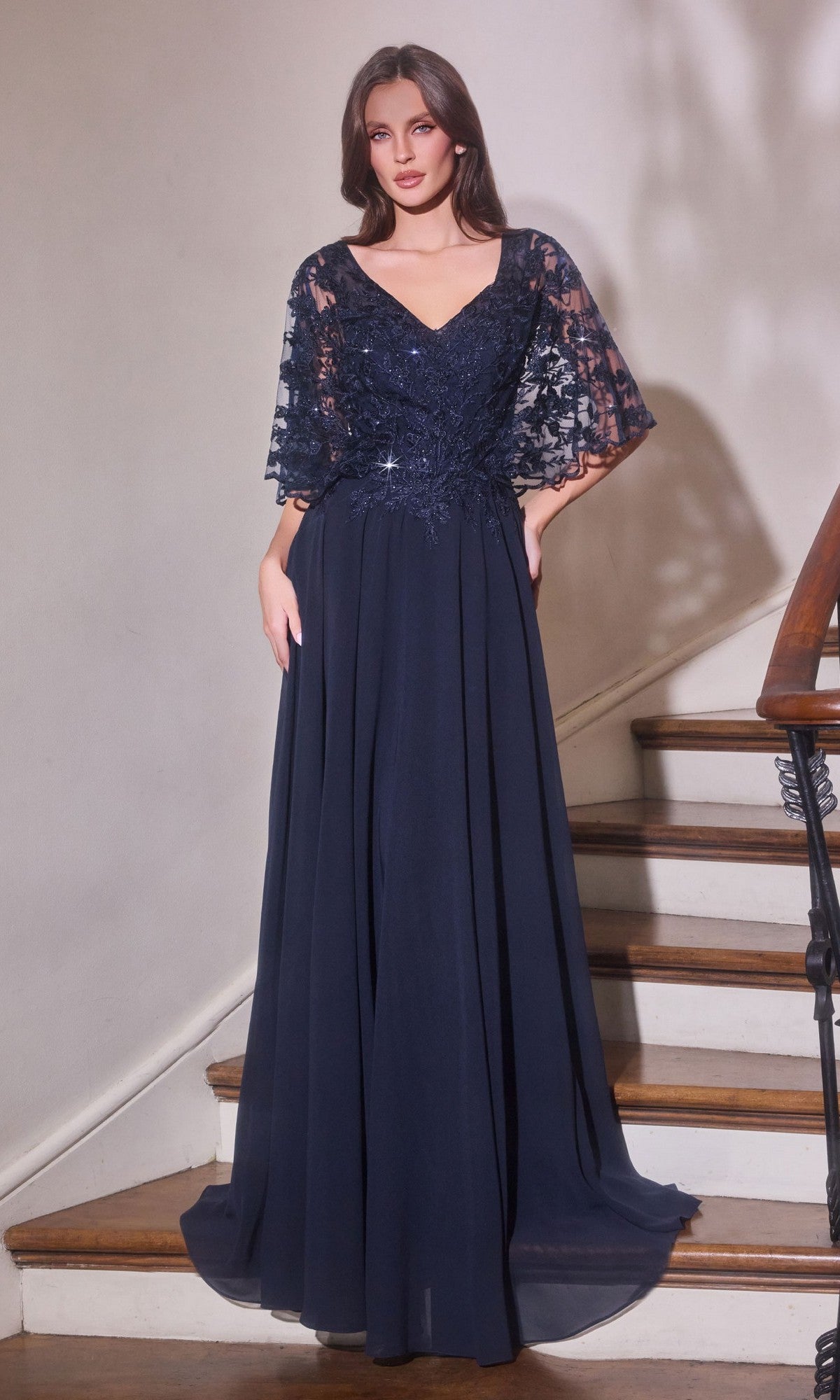A woman wears the Ladivine CL10 Long Formal Dress in navy, featuring a floor-length skirt, lace-embroidered bodice, and lace sleeves. She stands on a staircase facing forward with hands in the dress pockets.